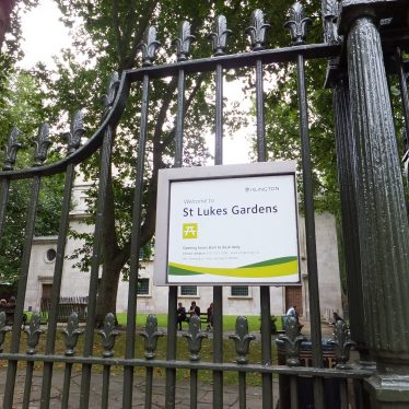 Railings surrounding a churchyard with trees and church behind and a notice about 'St Lukes Gardens Islington' | Louise Perkins