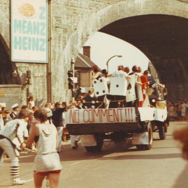 Float with gambling theme going under a bridge. Crowds including those in fancy dress. | Nuneaton Memories