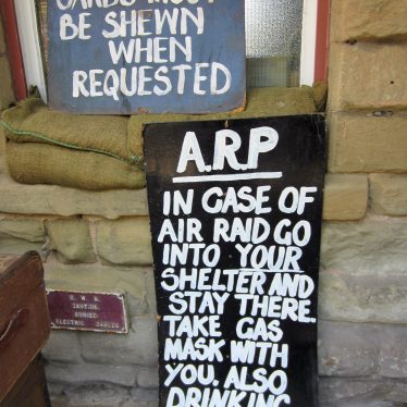 Sandbagged building with signs saying 'A.R.P. In case of air raid go into your shelter and stay there. Take gas mask with you. Also drinking water.' And 'Identity cards must be shewn when requested'. | Anne Langley