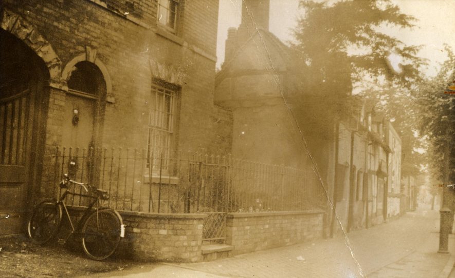 A sepia tinted photograph of a brick house with wide archway through, a bicycle in front and a row of timber-framed houses in the background | Warwickshire County Record Office reference CR 2487/Box 85/1224.