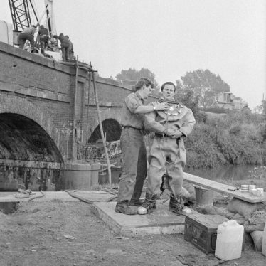 Diver standing in suit with helper in front of river bridge with crane and workmen | Warwickshire County Record Office PH 882/3124