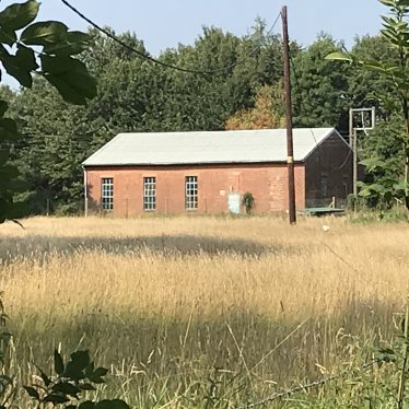 Long, low redbrick building with grey roof in field surrounded by trees | Image courtesy of Camille Newton