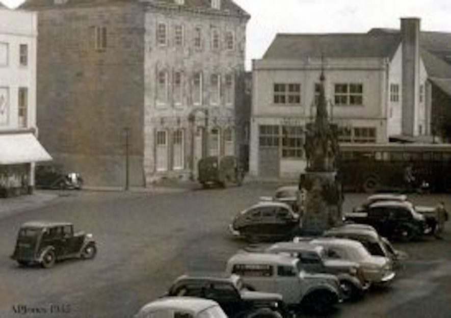 Black and white photo showing the side of Market Hall Museum, Fountain, edges of shops and old cars | Image courtesy of Alan Jones