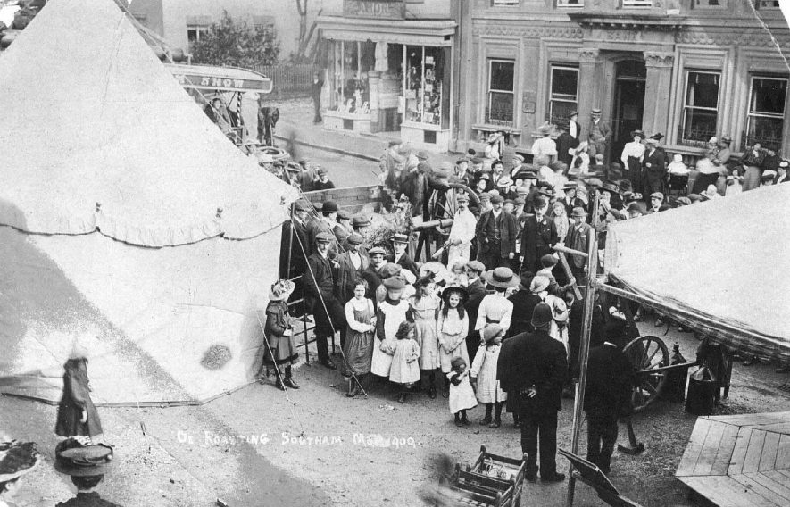 Ox roasting, Southam Mop 1909; shows a crowd gathered around the spit, amongst the tents and rides on Market Hill.  1909 |  IMAGE LOCATION: (Warwickshire County Record Office)