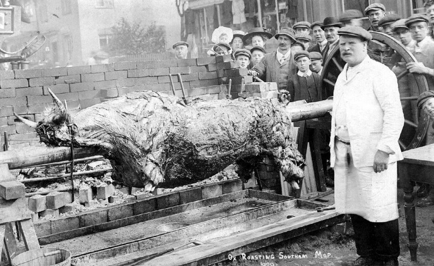 Ox roasting, Southam mop 1909; close up of the ox on the spit, with a crowd gathered behind it, one man holding a pitchfork. The man in charge of the ox roast is in the foreground.  1909 |  IMAGE LOCATION: (Warwickshire County Record Office)