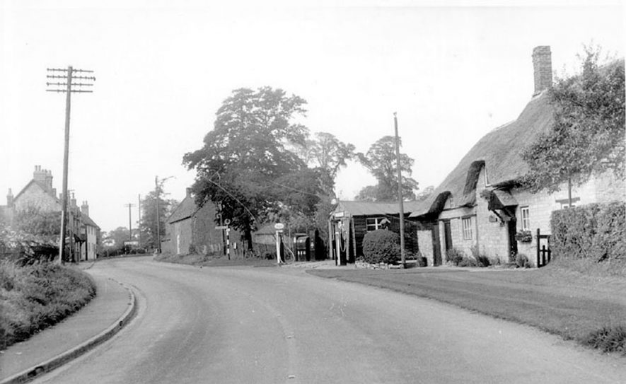 View of the Banbury Road in Ladbroke showing a stone built thatched cottage and a wooden garage.  1960s |  IMAGE LOCATION: (Warwickshire County Record Office)
