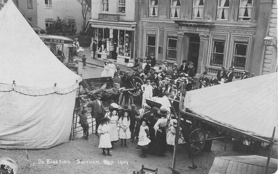 Ox roasting, Market Hill, Southam Mop 1909; view of tents, parts of rides, and crowd around the ox roast.  1909 |  IMAGE LOCATION: (Warwickshire County Record Office)
