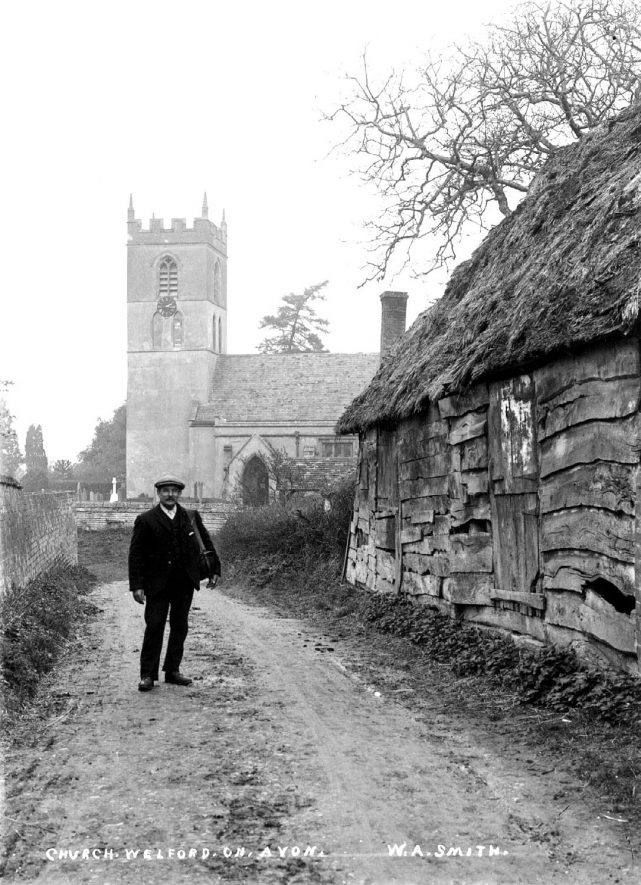 Man standing beside an old barn with church of St Peter in the background.  1900s |  IMAGE LOCATION: (Warwickshire County Record Office)