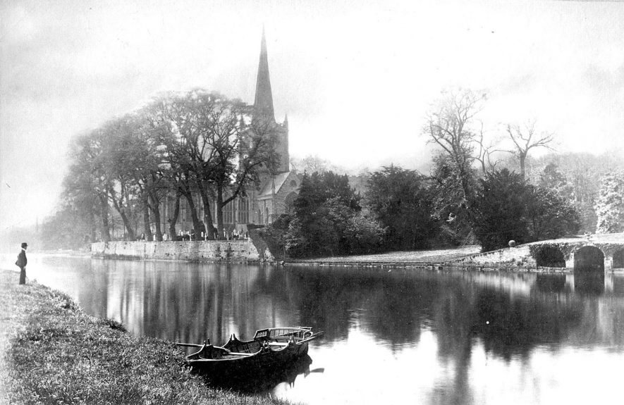 The parish church seen across the River Avon, with a man on the bank and a rowing boat moored by the bank, Stratford upon Avon.  1900s |  IMAGE LOCATION: (Warwickshire County Record Office)