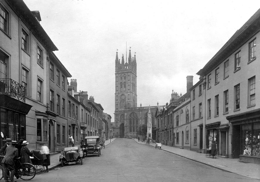 St Mary's Church tower from Church Street, Warwick. Hatter's shop and other shops. Terraced houses. Woman and girl with perambulator; two men with bicycle. Motor cars parked.  1920s |  IMAGE LOCATION: (Warwickshire County Record Office)
