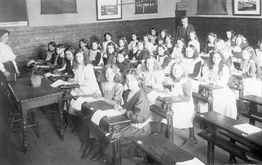 Interior of classroom at Abbey Street School, Nuneaton, showing girls at desks and two teachers, one man, one woman.  1900s |  IMAGE LOCATION: (Warwickshire County Record Office)
