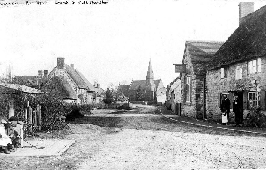 Looking south east to the Church, past the Post Office, and the Malt Shovel Inn. Post Office owner (?) with postman, child and bicycle outside the Post Office; three children sitting on opposite side of the road. Wooden wheelbarrow.  Postmark 1905 |  IMAGE LOCATION: (Warwickshire County Record Office)