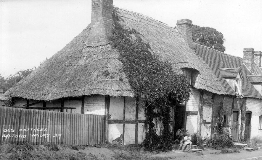 Children sitting on the step outside an old thatched, timber framed cottage. The cottage next door appears to be very small.  1920s |  IMAGE LOCATION: (Warwickshire County Record Office)