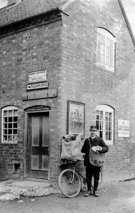 Long Itchington Post and Telegraph Office. Postman, possibly Mr A. Russell, standing outside with a bicycle and sacks of mail.  1910s |  IMAGE LOCATION: (Warwickshire County Record Office) PEOPLE IN PHOTO: Russell, A, Russell as a surname