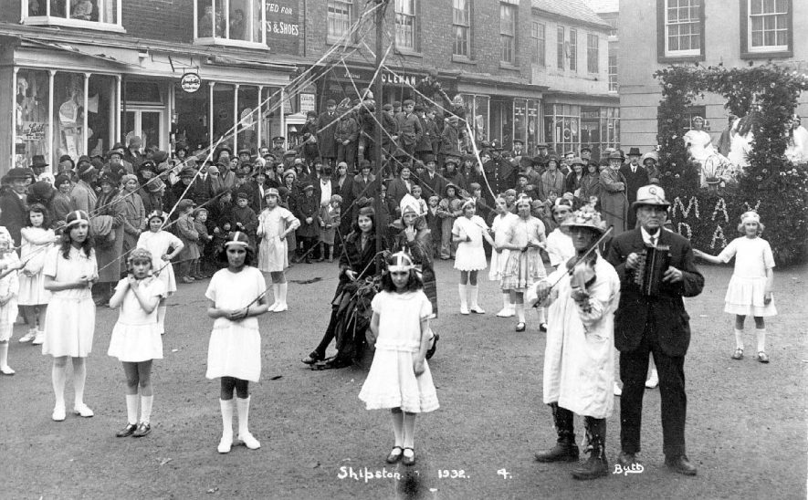 Children dressed for May Day celebrations with may pole in street, Shipston on Stour. Two musicians and watching crowd in front of shops. 1932 |  IMAGE LOCATION: (Warwickshire County Record Office)