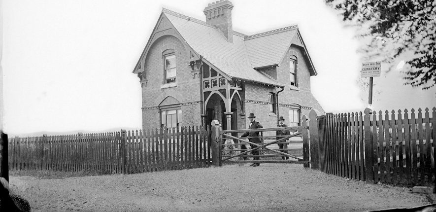The cemetery lodge, Nuneaton.  Two men in top hats standing by the gate, one of whom is carrying a spade.  1900s |  IMAGE LOCATION: (Warwickshire County Record Office)