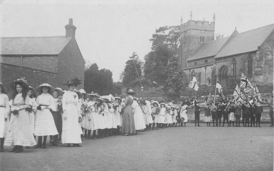 Mayday assembly in the school playground prior to parading round the village, Cubbington. Note the girls carrying flowers and the boys carrying flags. Circa 1910For more detailed information refer to - Cubbington by G.F.Peppitt. |  IMAGE LOCATION: (Warwickshire County Record Office)