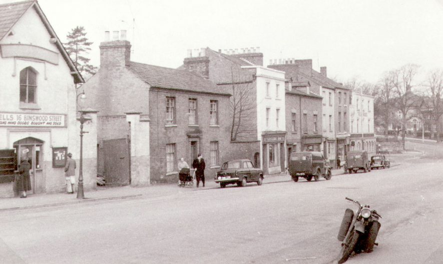 Shops and houses in Binswood Street, Leamington Spa.  1957  [Nos. 8 & 10 was a News agency with residential rooms,the name above the shop was E E Batchelder & Sons. The shop is behind the moving vehicle , as a two storey building with attic dormer windows.] |  IMAGE LOCATION: (Leamington Library)