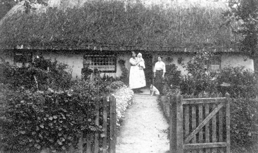 Bishops Tachbrook showing a thatched cottage and outside are Mrs Louise Bloomfield, the local midwife, the baby in her arms who later became Mrs Johnson and her mother, Mrs Emily Doughty.  1914 |  IMAGE LOCATION: (Warwickshire County Record Office) PEOPLE IN PHOTO: Johnson, Mrs, Johnson as a surname, Doughty, Mrs Emily, Doughty as a surname, Bloomfield, Mrs Louise, Bloomfield as a surname