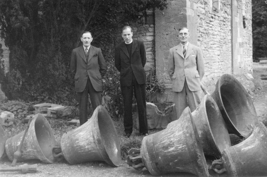 Three men, one the vicar, standing behind six church bells which are laying on the ground. Salford Priors.  1940s |  IMAGE LOCATION: (Warwickshire County Record Office)