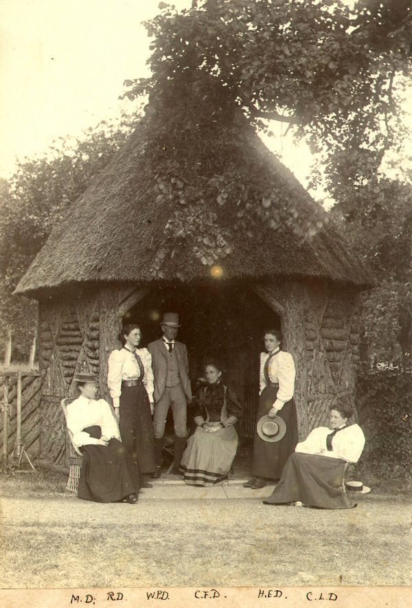 A Dickins family group standing in front of a small thatched summer house at Cherington. 1895.  From left to right - M. Dickins, R. Dickins, W.P. Dickins, C.F. Dickins, H.E. Dickins and C.L. Dickins. |  IMAGE LOCATION: (Warwickshire County Record Office) PEOPLE IN PHOTO: Dickins, W P, Dickins, R, Dickins, M, Dickins, H E, Dickins, C L, Dickins, C F, Dickins as a surname