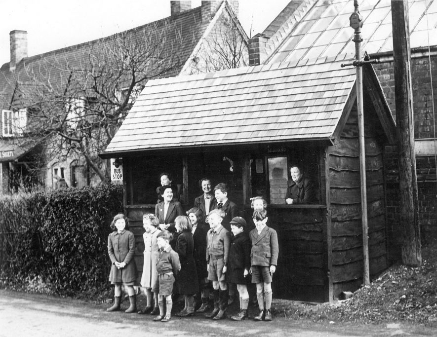 Offchurch bus shelter. From back left not known , Mrs. Hulme, Mrs Humphries, Mrs. Smith, Tony Boswell, Christine Owen, Elizabeth Jones, Chris Rose, Jean Jennings, Sheila Hawkes, Tony Jennings, David Owen, David Boswell.  1950s |  IMAGE LOCATION: (Warwickshire County Record Office) PEOPLE IN PHOTO: Smith, Mrs, Smith as a surname, Rose, Chris, Rose as a surname, Owen, David, Owen, Christine, Owen as a surname, Jones, Elizabeth, Jones as a surname, Jennings, Tony, Jennings, Jean, Jennings as a surname, Humphries, Mrs, Humphreys/Humphries as a surname, Hulme, Mrs, Hawkes, Sheila, Hawkes as a surname, Boswell, Tony, Boswell, David, Boswell as a surname