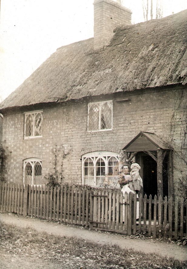 Thatched cottage with gothic style windows in Offchurch. Standing in front is a lady holding a small child.  1908
[The lady is Eliza Rose holding her son Bernard] |  IMAGE LOCATION: (Warwickshire County Record Office)