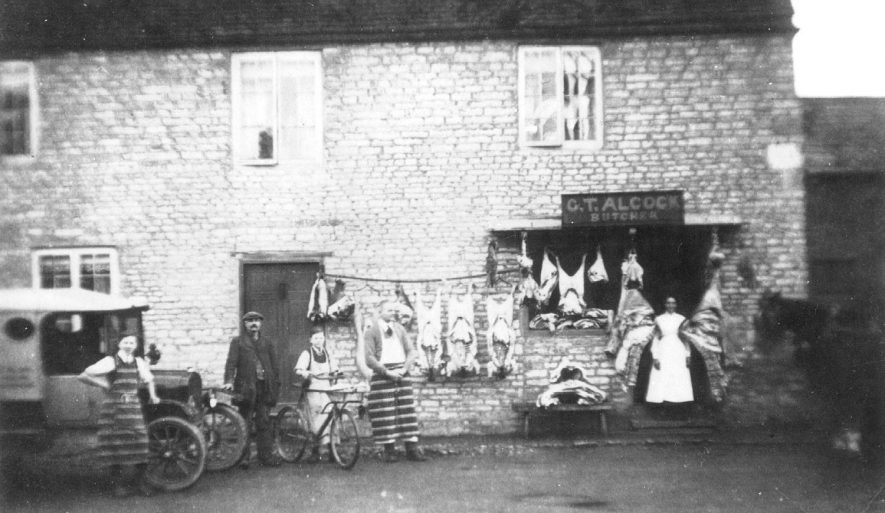 G T Alcock's butchers shop in Harbury. Carcasses are seen hanging on the outside wall and a delivery van and boy with cycle are also in the picture.  1900s |  IMAGE LOCATION: (Warwickshire Museums. Photographic Collections.) PEOPLE IN PHOTO: Dickins, Mr, Dickins as a surname
