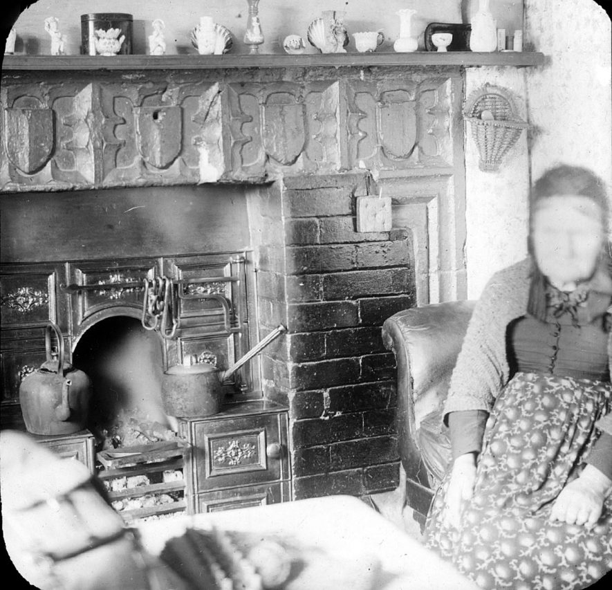 Fireplace in old rectory with carved coat of arms above.  Bricks and iron kitchener inserted into hearth.  Old woman seated.  Clifford Chambers.  1900 |  IMAGE LOCATION: (Warwickshire County Record Office)