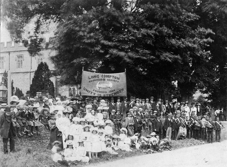 Gathering of the members of Long Compton Assurance Society in front of the church. They are holding a large banner aloft.  c.1904 |  IMAGE LOCATION: (Warwickshire County Record Office) IMAGE DATE: (c.1904)