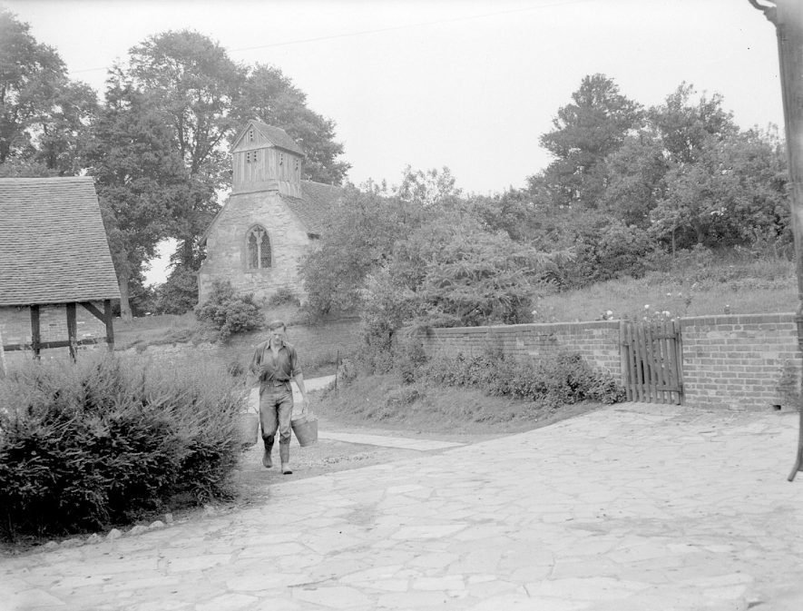 Holy Trinity church and old half timbered barn at Church Farm, Morton Bagot. The owner of the farm, Paul Sperryn is walking in the farm drive carrying two buckets. He farmed there between 1950 and 2004. July 20th 1956[ |  IMAGE LOCATION: (Warwickshire County Record Office)
