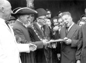 The mayoral party tasting the first slice of the pig roast at the Mop fair, Warwick. 1950s. A man with chain is handed a oplate by a smiling man with curly hair. |  IMAGE LOCATION: (Warwickshire County Record Office)