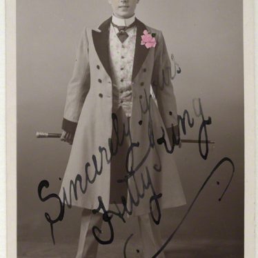Hetty King, a male impersonator who performed in Warwickshire. 1910s. Postcard published by Rotary Photographic Co Ltd. The subject is wearing a top hat, long coat, and is holding a cane behind her back. | Image originally from the National Portrait Gallery, London