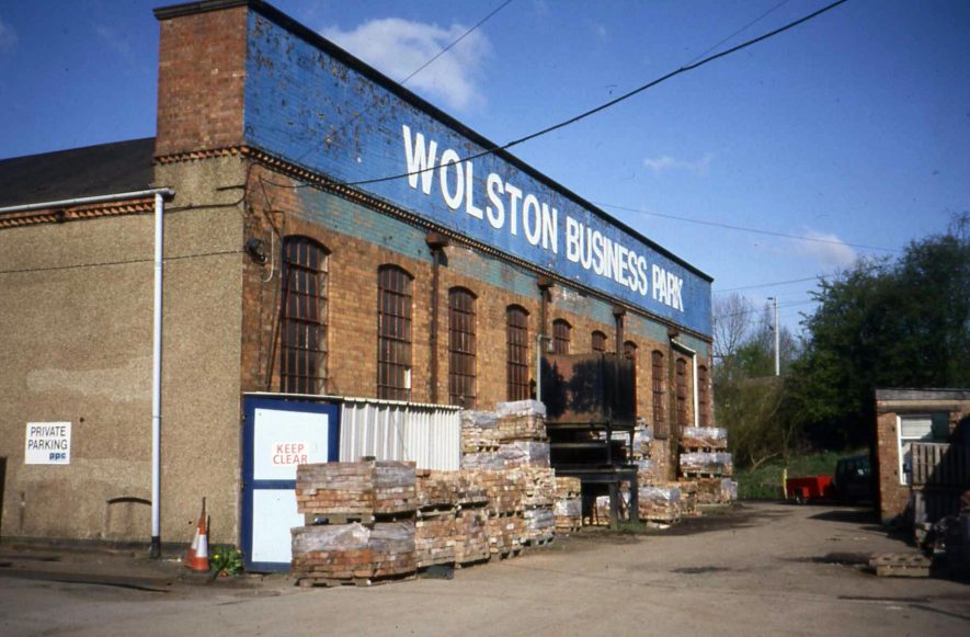 End of red brick factory building; blue painted top with 'Wolston Busines Park' written on in white. 10 metal windows; stacks of pallets outside and hut labelled 'keep clear'. | Image courtesy of Anne Langley
