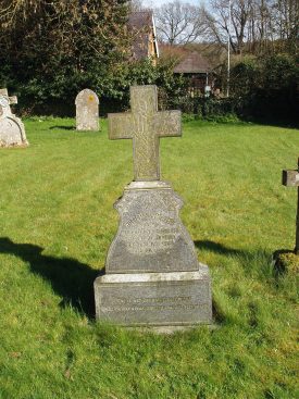 Cross over stone with inscription on in grassy churchyard. Other tombstones, trees and houses in the background | Image courtesy of Maureen Harris