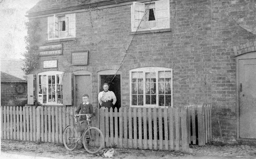 Church Lawford. Post Office and General Store, 1905. Probably Mrs. Rose Whiteman standing in the doorway of the house next to the Post Office whose husband, William Whiteman, was the Proprietor. Their son, Charlie, is standing with his bicycle by the fence. | Image courtesy of Pam Taylor
