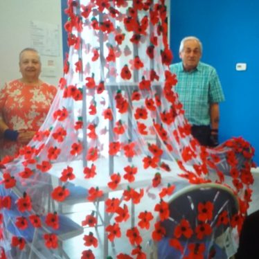 Lynne and Raymond stand beside the poppies that they made within the Rainbow Over-50s Club. This photo shows Lynne standing to the left of the frame, and Raymond standing to the right. In between them there are the poppies they made in their Over-50s Club all sewn onto sheer fabric which is suspended above them | Image Courtesy of Lynne Hodgson