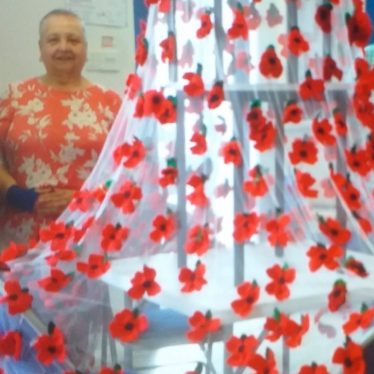 Lynne Hodgson stands beside the poppies that were made in the Over 50's Club. They are simple poppies made out of red and black felt, with four petals. In the photo they are woven onto netting which is suspended over some chairs. | Image Courtesy of Lynne Hogson