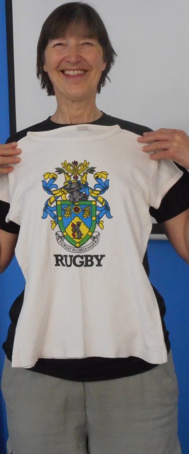 A tall brunette woman holds a white t-shirt with the Rugby Borough Crest, and the word RUGBY at the bottom against her. The crest has a knights head with a crown on top of it, with a traditional shield shape below it. The crest is decorated in blue, yellow, green and red. | Image Courtesy of Fern Hodges