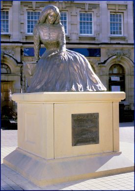 George Eliot Statue in Nuneaton, 2019. Metal statue of seated figure wearing a dress with a tight waist and full skirt, on a stone plinth with plaque attached; stone building behind | Image courtesy of John Morgan