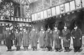 Brethren at Lord Leycester's Hospital, 1900s. Nine Brethren in gowns (with medals) and top hats in courtyard with gallery above, four shields with coats of arms and a porcupine | Warwickshire County Record Office reference PH352/187/39