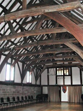 The Great Hall of Lord Leycester's Hospital, Warwick. Interior of hall showing fine timber roof springing from stone wall; timber framed partition at one end with shield with coat of arms; row of modern chairs | Image courtesy of Anne Langley