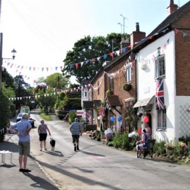 Bunting and flags; tea tables set outside cottages; a few people passing and a dog on a lead, 2020 | Roger Clemons