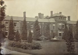 Brick-built building with slate roof; mostly 2-storey, but a central block is 3-storey with bay windows. Grounds in front with lawn, shrubs and trees | Warwickshire County Record Office reference CR 2941/21/2