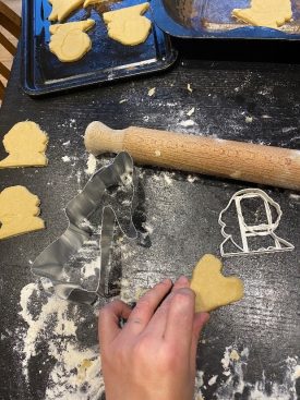 Colour photo, interior. dark wood table scattered with flour, dough, a rolling pin, biscuit cutters – in the shape of William Shakespeare- and cut out biscuits. One white hand reaches into the picture, holding a raw dough biscuit, in the shape of a love heart. 2021 | Image courtesy of Lady Kitt