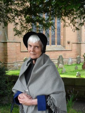 Dame at Lady Katherine Leveson Hospital in gown, shawl & bonnet. Woman in blue gown, black bonnet & black & white checked shawl standing in front of small crosses in graveyard with red sandstone Church behind | Image courtesy of Brita Wood