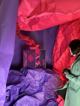 Christie, a woman with short black hair, is holding a camera and wearing a mask. She is looking at an artwork in an installation. The artwork is a cabinet that has been decoupaged with purple paper and pink paper cut artworks. It has hexagonal holes inside to hold scrolls that resemble the inside of a hive. The installation is a cave like structure made of textured large pieces of pink and purple paper. | Image courtesy of Christie Chan