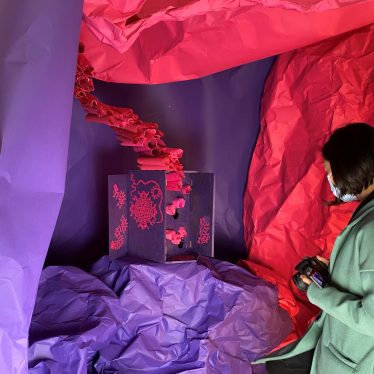 Christie, a woman with short black hair, is holding a camera and wearing a mask. She is looking at an artwork in an installation. The artwork is a cabinet that has been decoupaged with purple paper and pink paper cut artworks. It has hexagonal holes inside to hold scrolls that resemble the inside of a hive. The installation is a cave like structure made of textured large pieces of pink and purple paper. | Image courtesy of Christie Chan
