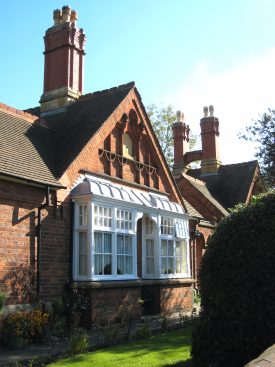 Redbrick 1-storey building with stone dressings, white-painted bay windows, tiled roof and tall chimneys; small front garden with lawn, shrubs and flowers | Image courtesy of Anne Langley