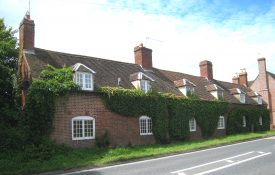 Coughton former almshouse. Red brick row with six white painted windows downstairs & six dormer window in a tiled roof with four chimneys; there are no doors, lots of creeper on the wall and a strip of grass fronting onto a road | Image courtesy of Anne Langley